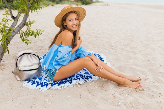 Elegante hermosa mujer sonriente sentada en la arena con las piernas delgadas en vacaciones de verano en la playa tropical con sombrero de paja, mochila plateada