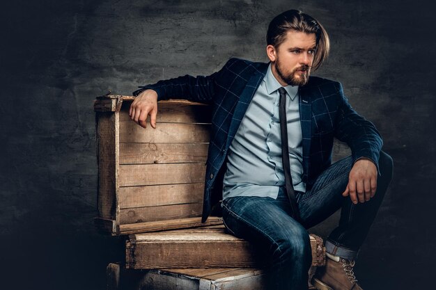 El elegante hipster barbudo se sienta en una caja de madera en un estudio sobre un fondo gris oscuro.