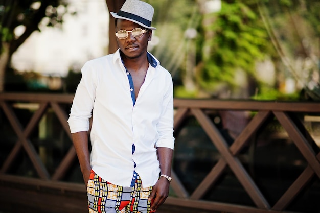 Elegante hombre afroamericano con camisa blanca y pantalones de colores con sombrero y gafas posó al aire libre Chico modelo de moda negro