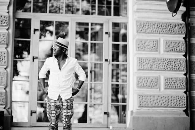 Elegante hombre afroamericano con camisa blanca y pantalones de colores con sombrero y gafas posó al aire libre Chico modelo de moda negro