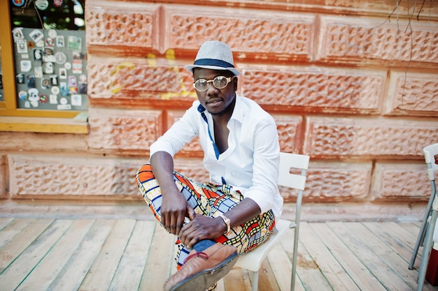 Elegante hombre afroamericano con camisa blanca y pantalones de colores con sombrero y gafas posó al aire libre Chico modelo de moda negro