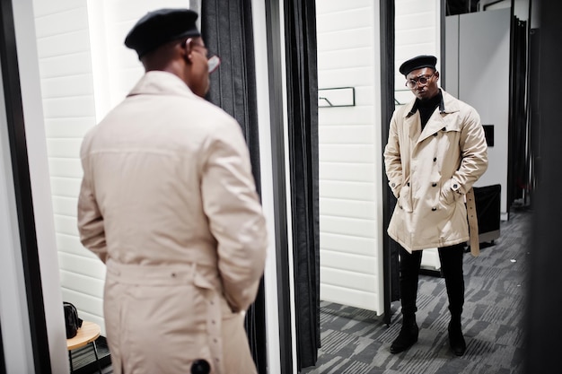 Elegante hombre afroamericano casual con boina negra y gabardina blanca en la tienda de ropa de la sala de montaje mirando al espejo