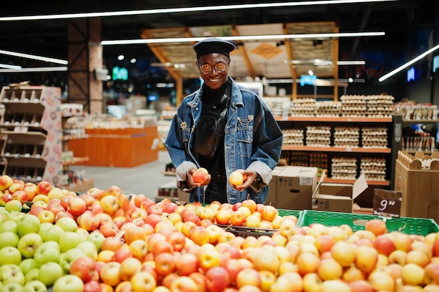 Elegante hombre afroamericano casual con chaqueta de jeans y boina negra revisando frutas de manzana en la sección orgánica del supermercado