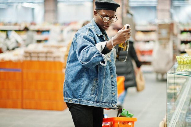 Elegante hombre afroamericano casual con chaqueta de jeans y boina negra sosteniendo una canasta y mirando una botella de vino comprando en el supermercado