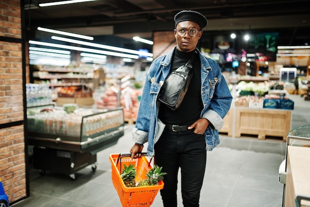 Elegante hombre afroamericano casual con chaqueta de jeans y boina negra sosteniendo una canasta con piñas en la sección orgánica de frutas del supermercado