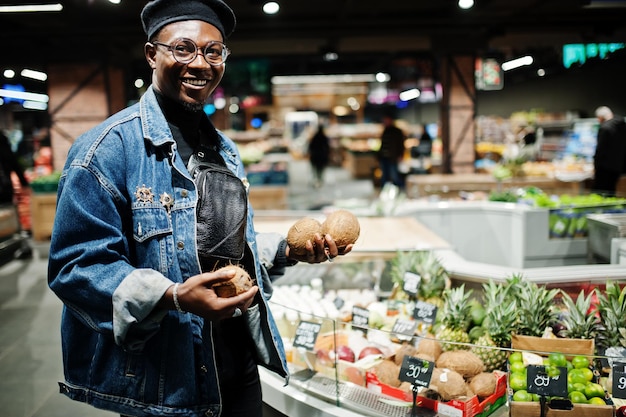 Elegante hombre afroamericano casual con chaqueta de jeans y boina negra sosteniendo cocos en la sección orgánica de frutas del supermercado