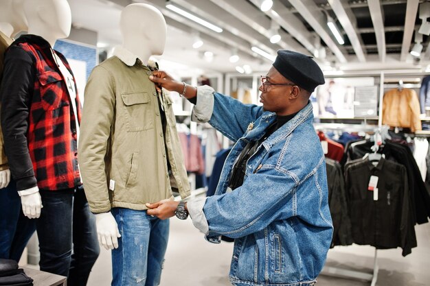 Elegante hombre afroamericano casual con chaqueta de jeans y boina negra en la tienda de ropa mirando una nueva chaqueta en maniquí