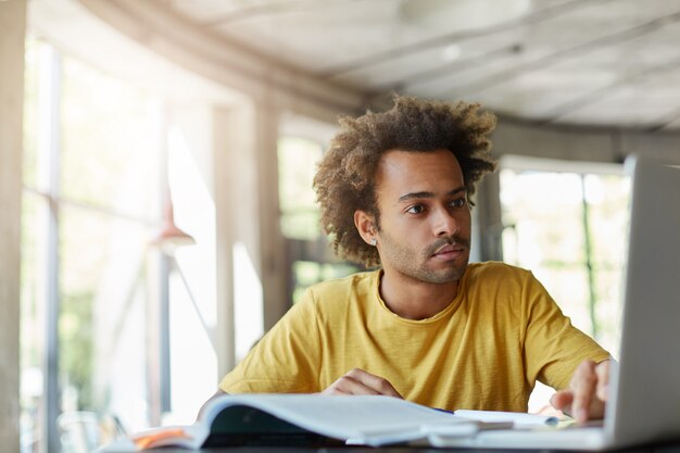 Elegante hombre afroamericano hipster con peinado tupido con camiseta casual que se enfoca en la pantalla de la computadora portátil sentado en una espaciosa habitación luminosa con grandes ventanales que trabaja con literatura e internet