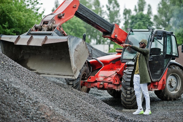 Elegante hombre afroamericano con sombrero y gafas de sol posó al aire libre bajo la lluvia contra el tractor con un balde