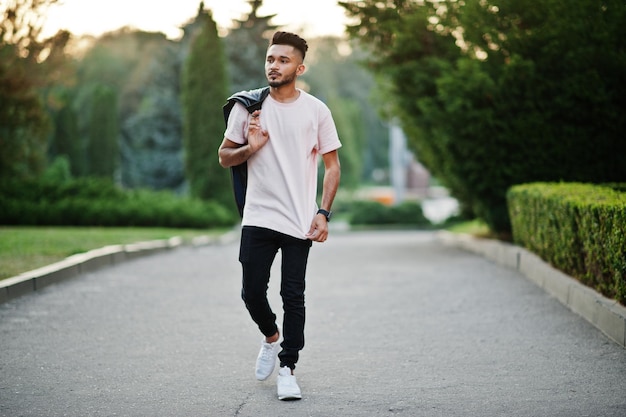 Elegante hombre de barba india en camiseta rosa con chaqueta de cuero negro en el hombro Modelo de India caminando en la ciudad de la tarde