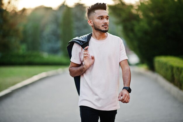 Elegante hombre de barba india en camiseta rosa con chaqueta de cuero negro en el hombro Modelo de India caminando en la ciudad de la tarde