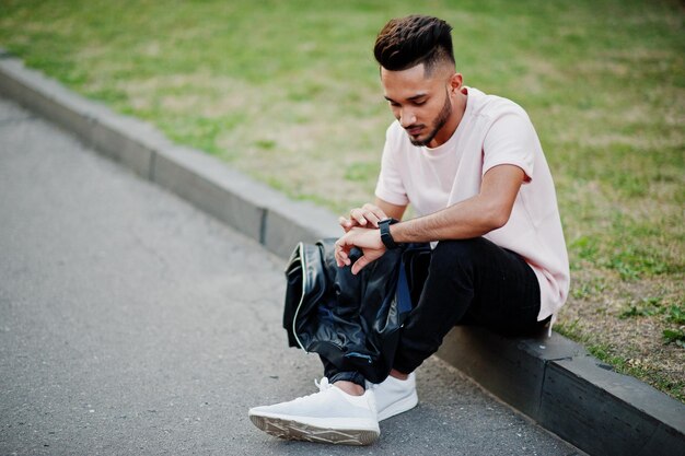 Elegante hombre de barba india con camiseta rosa Modelo de India sentado en el jardín verde de la ciudad y mirando sus relojes modernos