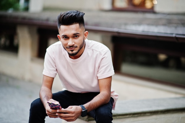 Elegante hombre de barba india con camiseta rosa Modelo de India sentado con el teléfono móvil en las manos al aire libre en las calles de la ciudad