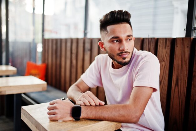 Elegante hombre de barba india con camiseta rosa sentado contra una mesa de madera de café India modelo posado al aire libre