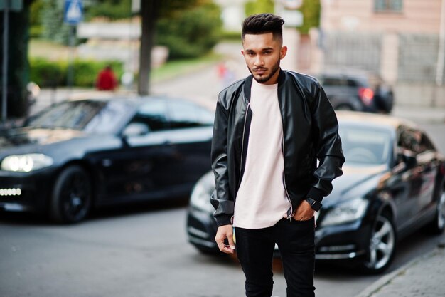 Elegante hombre de barba india con chaqueta de cuero negro contra el coche Modelo de India posado en las calles de la ciudad