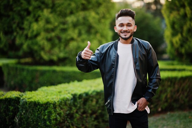 Elegante hombre de barba india con chaqueta de cuero negro muestra el pulgar hacia arriba Modelo de India posado en el jardín verde de la ciudad