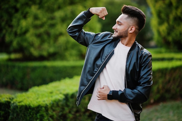 Elegante hombre de barba india con chaqueta de cuero negro muestra su mano muscular Modelo de India posado en el jardín verde de la ciudad