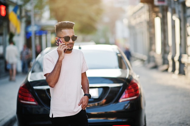 Elegante hombre de barba india con gafas de sol y camiseta rosa contra un automóvil de lujo y hablando por teléfono móvil Modelo rico de India posado al aire libre en las calles de la ciudad