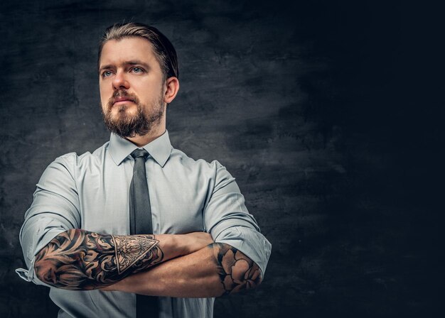 Elegante hombre barbudo con brazos tatuados, vestido con una camisa posando sobre fondo gris.