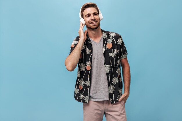 Elegante hombre barbudo con camiseta gris estampada y plantas beige mirando hacia otro lado y posando con auriculares blancos sobre fondo azul