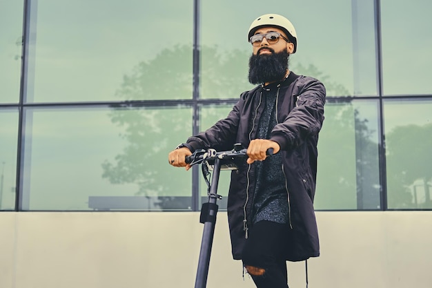 Elegante hombre barbudo con gafas de sol posando en scooter eléctrico sobre un moderno edificio de fondo.