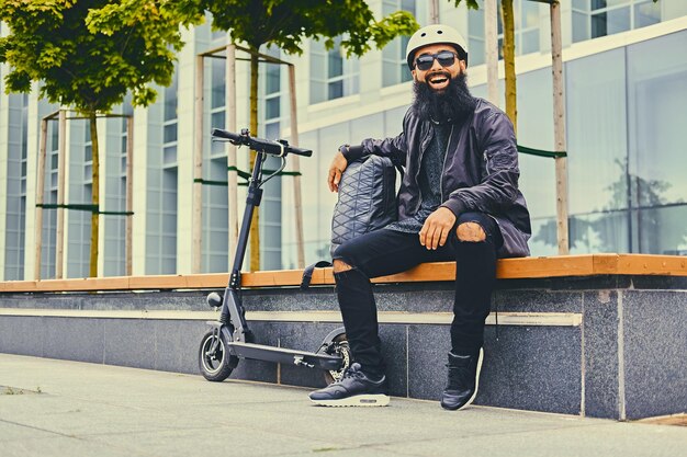 Elegante hombre barbudo con gafas de sol relajándose en un banco después de andar en scooter eléctrico en el centro.