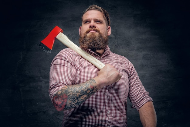 Elegante hombre barbudo con tatuajes en los brazos con camisa a cuadros rosa y sostiene un hacha.