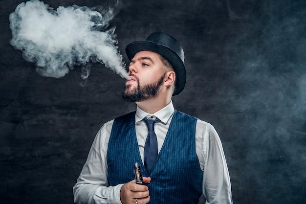 Un elegante hombre barbudo vestido con chaleco y sombrero de copa fumando un cigarrillo electrónico.