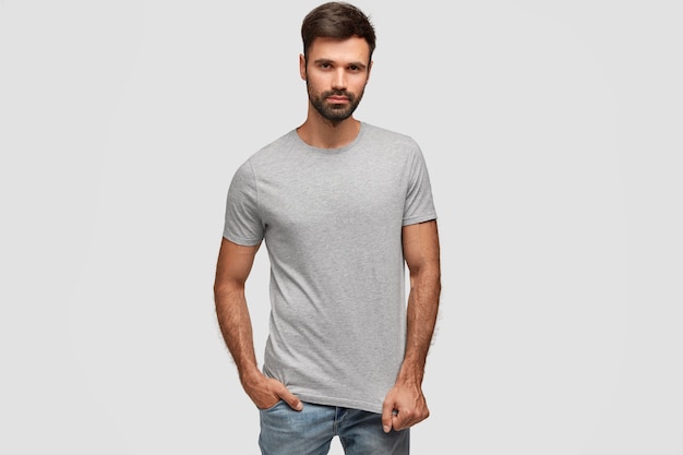 Elegante hombre guapo con barba y barba densa y oscura, vestido con camiseta casual, mantiene la mano en el bolsillo de los pantalones vaqueros