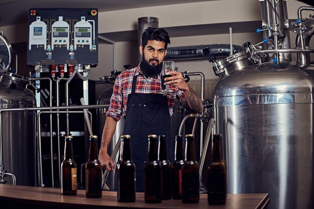 Elegante hombre indio barbudo con una camisa de lana y un delantal sostiene un vaso de cerveza, parado detrás del mostrador en una cervecería.