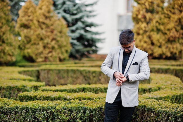 Elegante hombre indio con bindi en la frente y anteojos en traje posado al aire libre contra arbustos verdes en el parque y mirando sus relojes