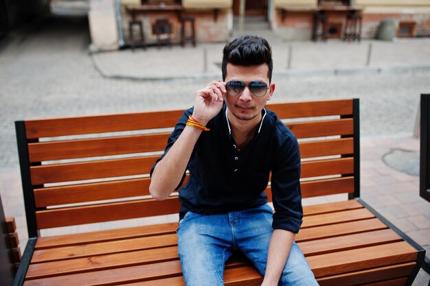 Elegante hombre modelo indio con ropa informal, camisa negra y gafas de sol, posado al aire libre en la calle de la India y sentado en un banco