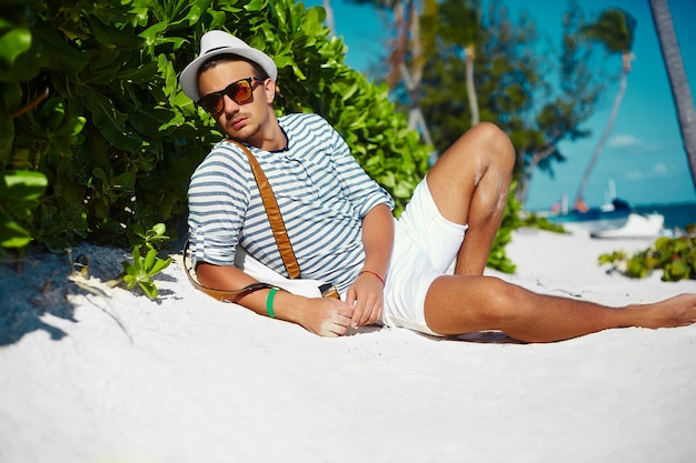 Elegante hombre modelo masculino joven tumbado en la arena de la playa con sombrero de verano hipster