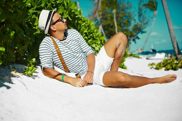 Elegante hombre modelo masculino joven tumbado en la arena de la playa con sombrero de verano hipster