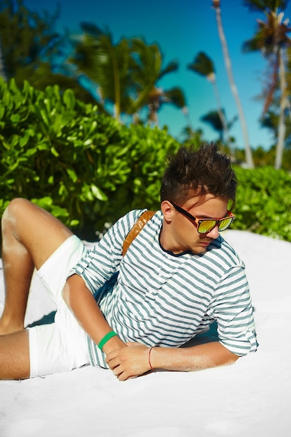 Elegante hombre modelo masculino joven tumbado en la arena de la playa con sombrero de verano hipster