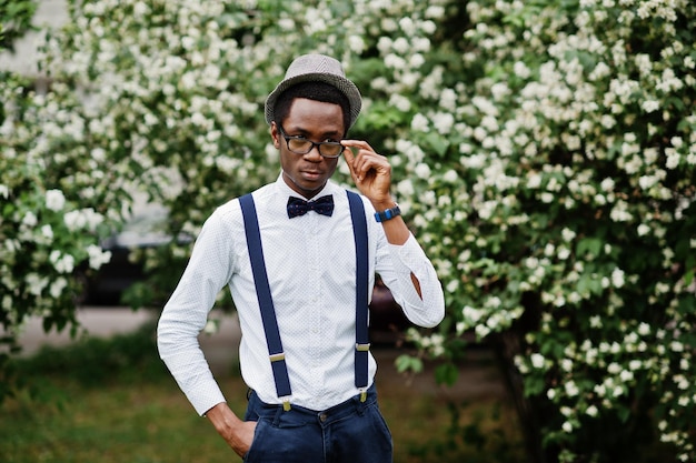 Elegante hombre de negocios afroamericano en pantalones con tirantes y camisa con sombrero de pajarita y gafas posadas al aire libre