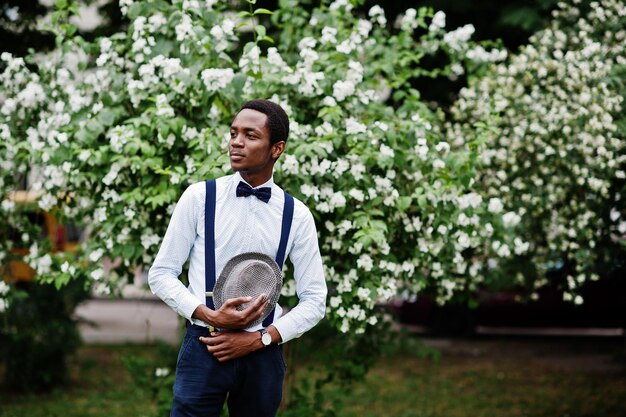 Elegante hombre de negocios afroamericano en pantalones con tirantes y camisa con sombrero de pajarita posado al aire libre