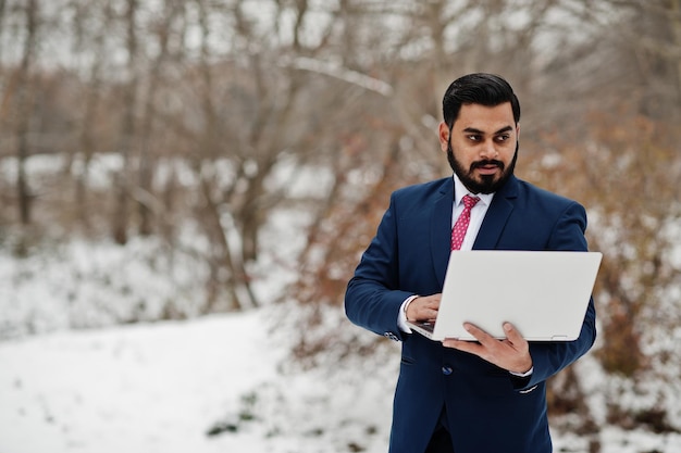 Foto gratuita elegante hombre de negocios de barba india en traje posado en el día de invierno al aire libre con una computadora portátil en las manos