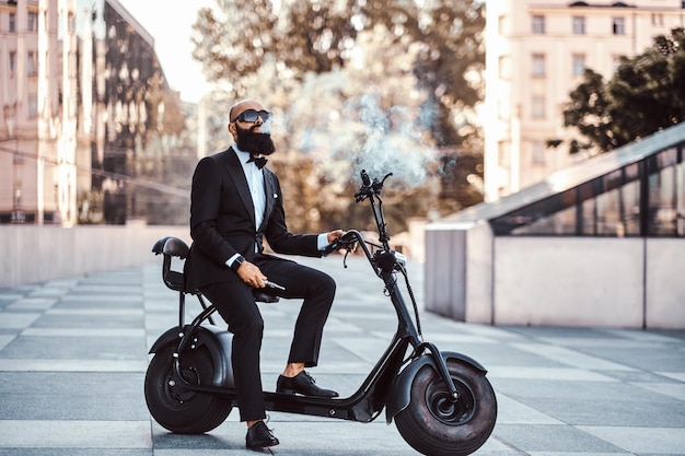 Elegante hombre de negocios calvo con gafas de sol está fumando vaporizador mientras está sentado en su bicicleta eléctrica.