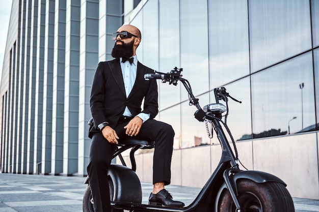 Foto gratuita un elegante hombre de negocios calvo con gafas de sol está sentado en su scooter eléctrico cerca de un edificio de cristal.