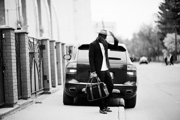 Elegante hombre negro con gafas con sombrero en traje con bolso contra coche de lujo Rico empresario afroamericano