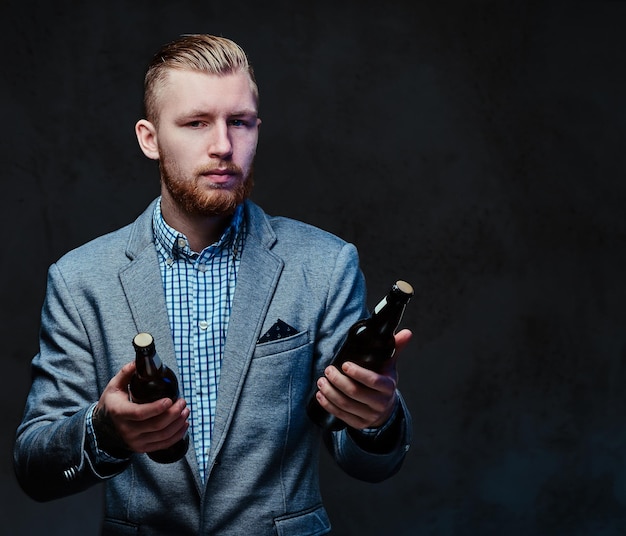 Elegante hombre pelirrojo con barba vestido con traje sostiene una botella con cerveza artesanal sobre fondo gris.