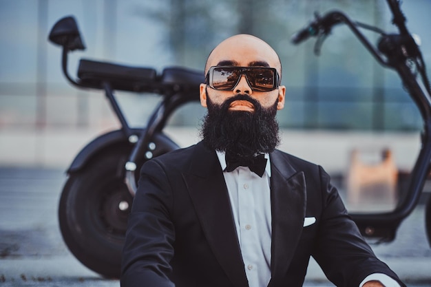 Foto gratuita elegante hombre pensativo con gafas de sol está sentado en las escaleras cerca de su scooter eléctrico.