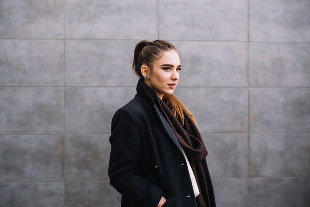 Elegante joven en abrigo con bufanda cerca de pared gris