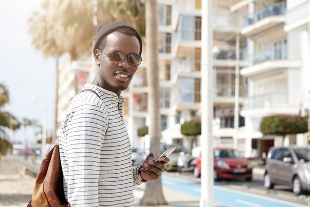 Elegante joven afroamericano en tonos y sombrero que busca ubicaciones a través de aplicaciones en línea para viajes o navegación gps, usando 3g y 4g en el teléfono móvil mientras camina en metrópolis extranjera