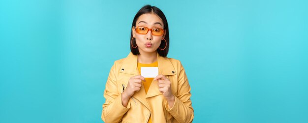 Elegante joven asiática con gafas de sol mostrando tarjeta de crédito y sonriendo recomendando pagos bancarios sin contacto o descuentos en la tienda de pie sobre fondo azul.