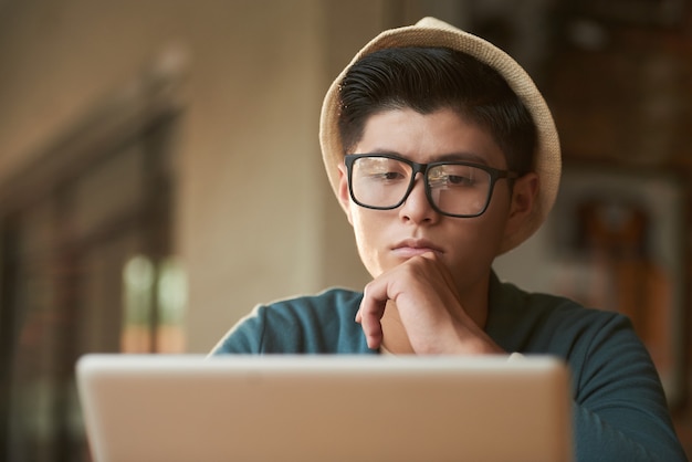 Elegante joven asiática con sombrero y gafas sentado en la cafetería y mirando la pantalla