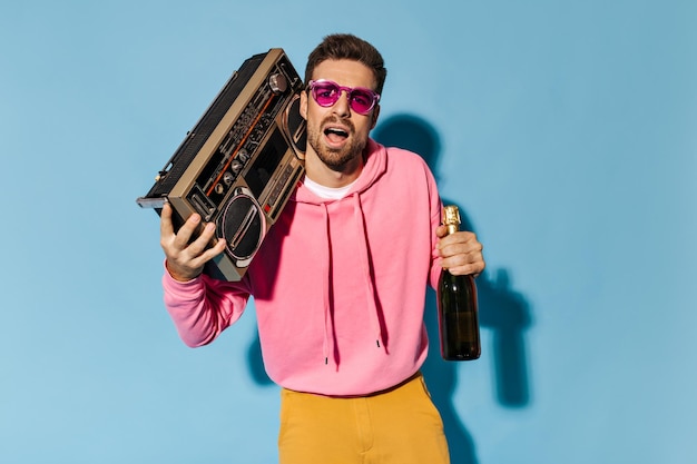 Elegante joven barbudo con gafas de sol rosas y capucha sostiene un tocadiscos retro y una botella de champán sobre fondo azul.