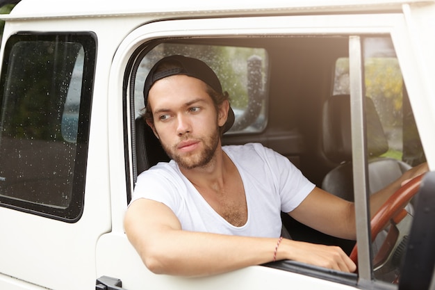 Elegante joven barbudo hipster en camiseta y snapback tirando de su jeep blanco después de ser detenido por la policía en un camino rural. Hombre guapo disfrutando de viaje por carretera