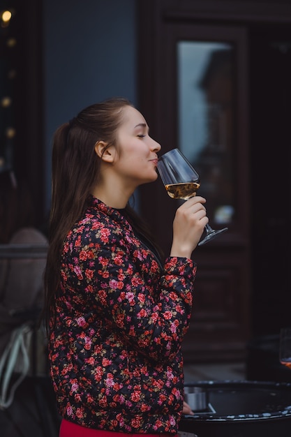 Elegante joven bebe vino en un café de la calle en una terraza de verano. Una muchacha con pelo largo goza de una copa de vino en una tarde del verano. Retrato. de cerca